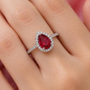 Pode incluir: Um anel com uma pedra vermelha em forma de pera, rodeada por um halo de pequenas pedras brancas numa banda de metal prateado. A pedra central em forma de lágrima está engastada com uma borda delicada de pedras brancas brilhantes.