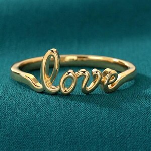 May include: A gold ring spelling out the word "love" in a cursive font. The ring is displayed on a teal background. The ring is a piece of jewellery.