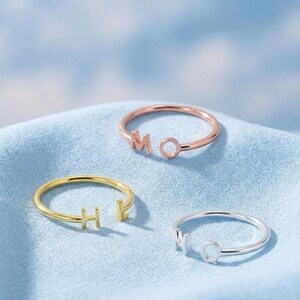 May include: Three open-style rings featuring individual letter charms. One ring is rose gold with the letters M and O, one is gold with the letters H and E, and one is silver with the letters H and Q. Each ring has a thin, circular band design.