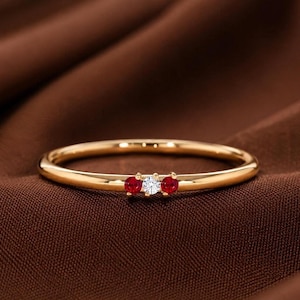 May include: A thin gold band ring featuring three small, round-cut stones set in a horizontal row. The centre stone is clear and colourless, flanked on each side by a vibrant, deep red stone. The metal has a smooth, polished, reflective finish.