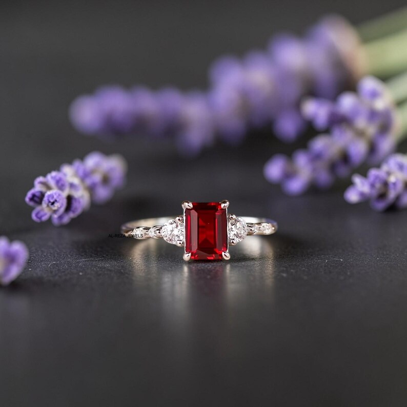 May include: A silver ring with a rectangular, deep red gemstone as the centerpiece, flanked by two smaller, round, white gemstones. The ring is set against a dark background with blurred purple flowers.