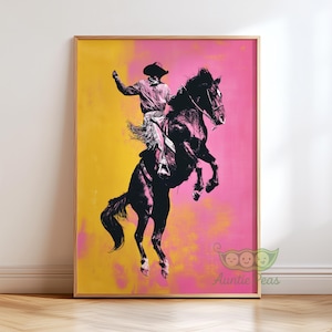 May include: Framed art print featuring a cowboy riding a rearing horse. The artwork is rendered in shades of black and pink against a yellow and pink background. The cowboy is wearing a hat and fringed chaps. The print is in a wooden frame.