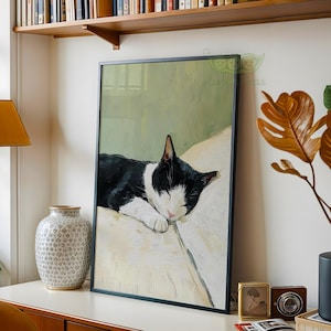 May include: Framed art print featuring a sleeping black and white cat. The cat is resting on a white surface against a light green background. The artwork is displayed in a black frame and is a charming addition to any home decor.