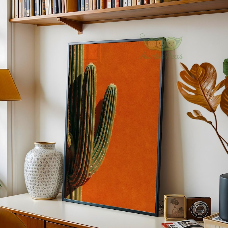 May include: Framed art print featuring a close-up of a saguaro cactus against a vibrant orange background. The cactus is green with vertical ridges and spines. The artwork is in a black frame, suitable for home decor.
