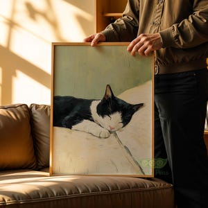 May include: A framed art print featuring a sleeping black and white cat. The cat is resting on a light-colored surface against a pale green background. The artwork has a soft, painterly style, and is held by a person.