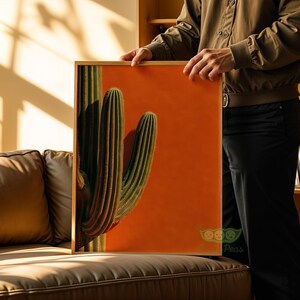 May include: A framed art print featuring a vibrant cactus against a solid orange background. The cactus is a deep green with vertical ridges, and the frame is a natural wood tone. The artwork is held by a person.