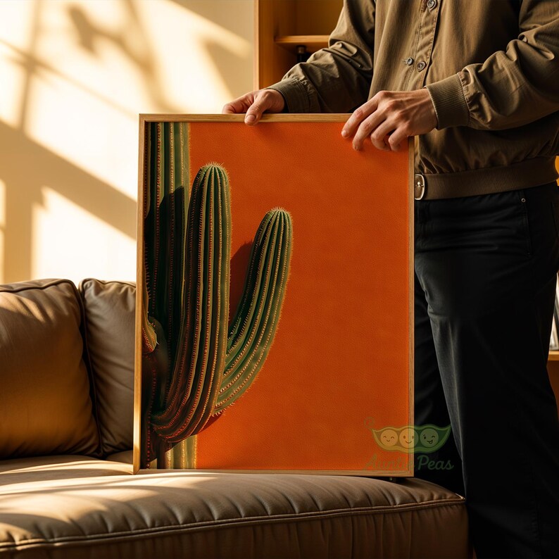 May include: A framed art print featuring a vibrant cactus against a solid orange background. The cactus is a deep green with vertical ridges, and the frame is a natural wood tone. The artwork is held by a person.