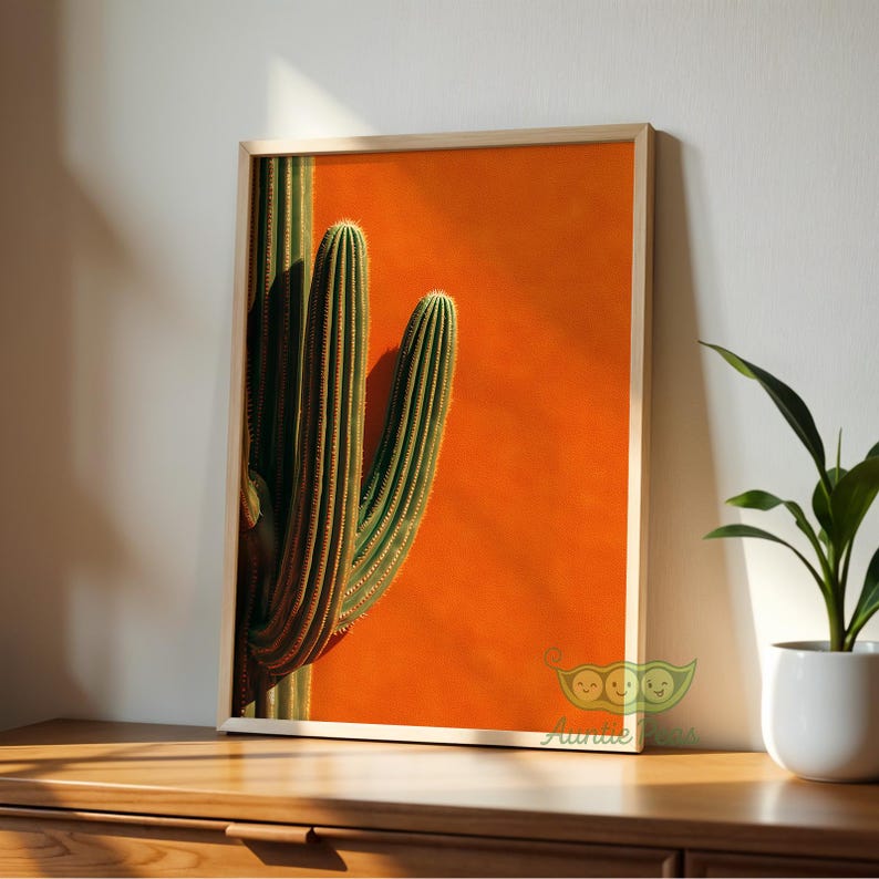 May include: Framed art print featuring a vibrant cactus against a bright orange background. The cactus is green with vertical ridges, and the frame is a light wood color. The artwork is displayed on a wooden surface.