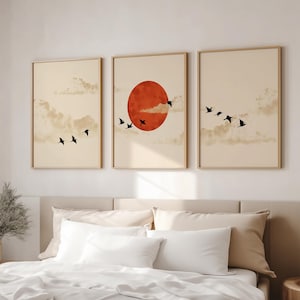 May include: Three framed art prints with a minimalist design. Each print has a beige background with clouds and black birds in flight. The central print features a large red sun. The frames are a light wood colour.