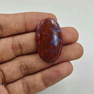 Könnte beinhalten: Ein ovaler Cabochon-Edelstein mit einer polierten Oberfläche, einer tief rotbraunen Grundfarbe und komplizierten, wirbelnden hellblauen und lavendelfarbenen Adern, die sich durch den Stein ziehen und ein glattes, glänzendes Finish aufweisen.