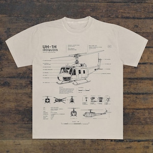 Könnte beinhalten: Ein beiges T-Shirt mit einer schwarzen technischen Strichzeichnung eines UH-1H Iroquois Mehrzweckhubschraubers. Die Grafik enthält detaillierte Schaltpläne, Beschriftungen und technische Daten des Fluggeräts im Stil eines Bauplans.