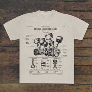 May include: A beige t-shirt featuring a black technical diagram of an internal combustion engine, piston, and crankshaft assembly. The graphic includes detailed labels, cross-section views, and technical specifications printed in a clean, sans-serif font.