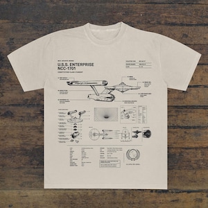 May include: A beige short-sleeve t-shirt featuring a black technical blueprint graphic of the U.S.S. Enterprise NCC-1701. The design includes detailed schematics, labels, and technical specifications for the Constitution-class starship.