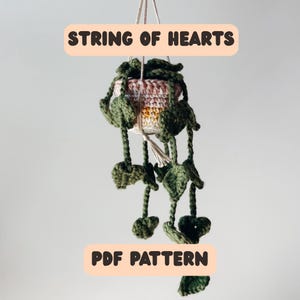 Op de afbeelding: Een gehaakte hangende plantenpot met een String of Hearts plant. De pot is een mix van roze, wit en geel. De plant heeft groene hartvormige bladeren. De tekst "STRING OF HEARTS" en "PDF PATTERN" worden weergegeven.