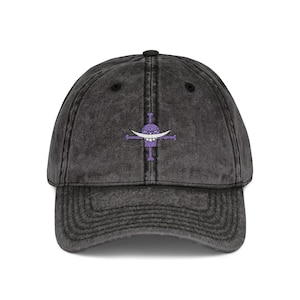 Op de afbeelding: Een donkergrijze baseballcap met een vintage wassing. De cap heeft een paars geborduurd embleem van een gestileerde witte snor en een paars kruisontwerp. De cap heeft een gebogen klep en een knoop aan de bovenkant.