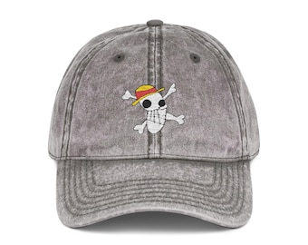 May include: Gray baseball cap with a distressed finish. The cap features an embroidered skull and crossbones logo wearing a yellow and red straw hat. The hat has a curved brim and an adjustable strap for a comfortable fit.