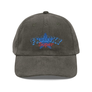 Puede incluir: Gorra de béisbol de pana verde oliva con la palabra "FRANKY" en azul y "Super" en rojo, bordada sobre una estrella azul. La gorra tiene una visera curva y un cierre de correa de tela.