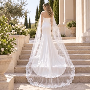 May include: A strapless white floor-length gown with a fitted bodice and a flared skirt, paired with a long, sheer, floor-sweeping veil featuring intricate white lace floral embroidery along the edges, cascading down the back of the garment.