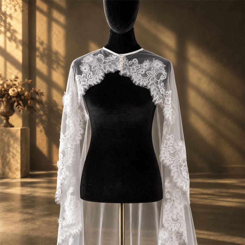 May include: A white sheer tulle bridal capelet featuring intricate floral lace trim along the edges and a high neckline with a small button closure. The delicate fabric drapes over the shoulders, showcasing detailed embroidery and scalloped borders.