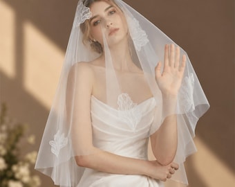May include: A sheer white tulle wedding veil featuring delicate white floral embroidery along the edges and scattered across the fabric. The veil is draped over the head, showcasing a soft, lightweight texture and intricate stitched leaf patterns.