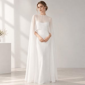May include: A floor-length white gown featuring a sleeveless bodice with a scoop neckline and a sheer, flowing cape overlay that extends from the shoulders to the floor, creating a layered silhouette with a clean, solid white finish throughout.