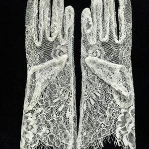 May include: A pair of off-white lace gloves featuring a detailed floral pattern throughout the fabric. The gloves extend to the wrist and are finished with a delicate, fringed scalloped edge at the opening, showcasing intricate needlework.
