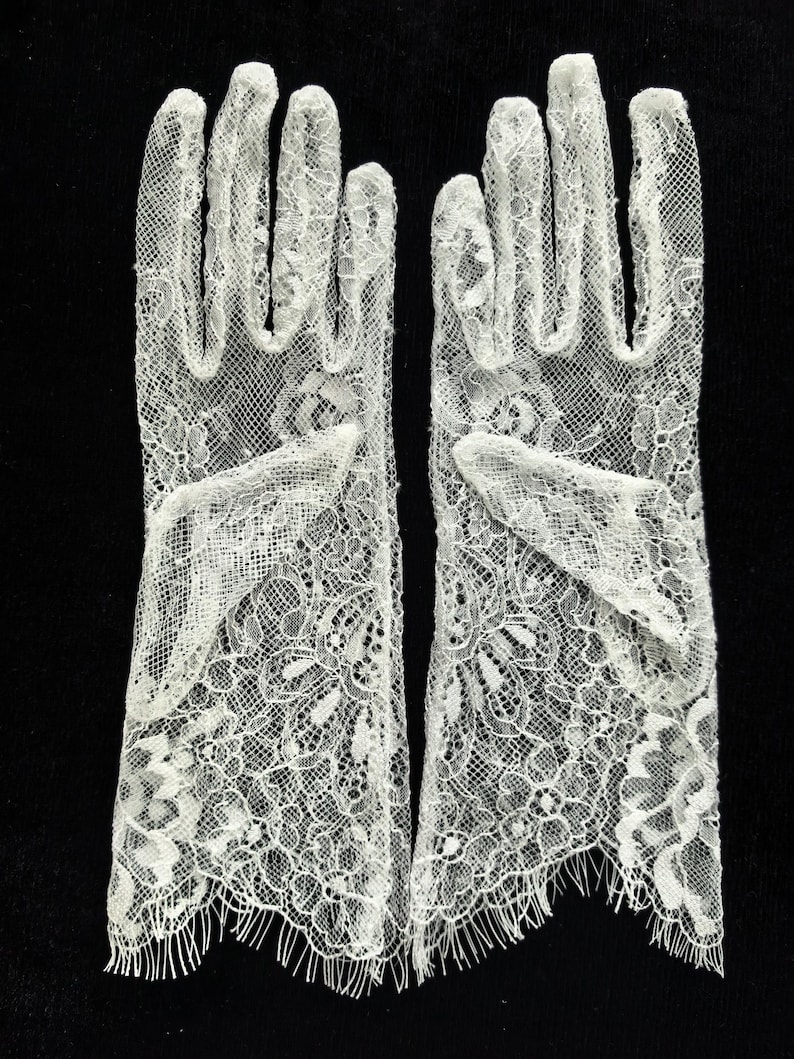 May include: A pair of off-white lace gloves featuring a detailed floral pattern throughout the fabric. The gloves extend to the wrist and are finished with a delicate, fringed scalloped edge at the opening, showcasing intricate needlework.