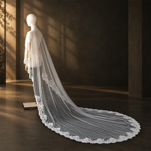 May include: A long, sheer white bridal veil featuring intricate floral lace embroidery along the edges. The veil drapes from the shoulders of a mannequin, extending onto the floor in a sweeping train with a scalloped lace border design.