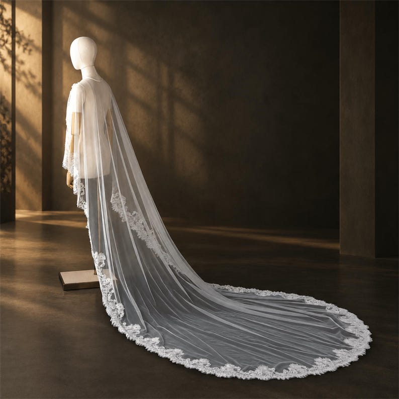 May include: A long, sheer white bridal veil featuring intricate floral lace embroidery along the edges. The veil drapes from the shoulders of a mannequin, extending onto the floor in a sweeping train with a scalloped lace border design.