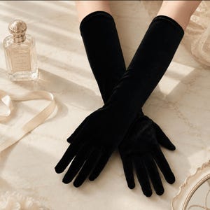 May include: A pair of long, black velvet opera gloves that extend past the wrist. The gloves feature a smooth, soft-textured fabric with a deep black color and a fitted design that covers the fingers and forearms, creating a sleek appearance.