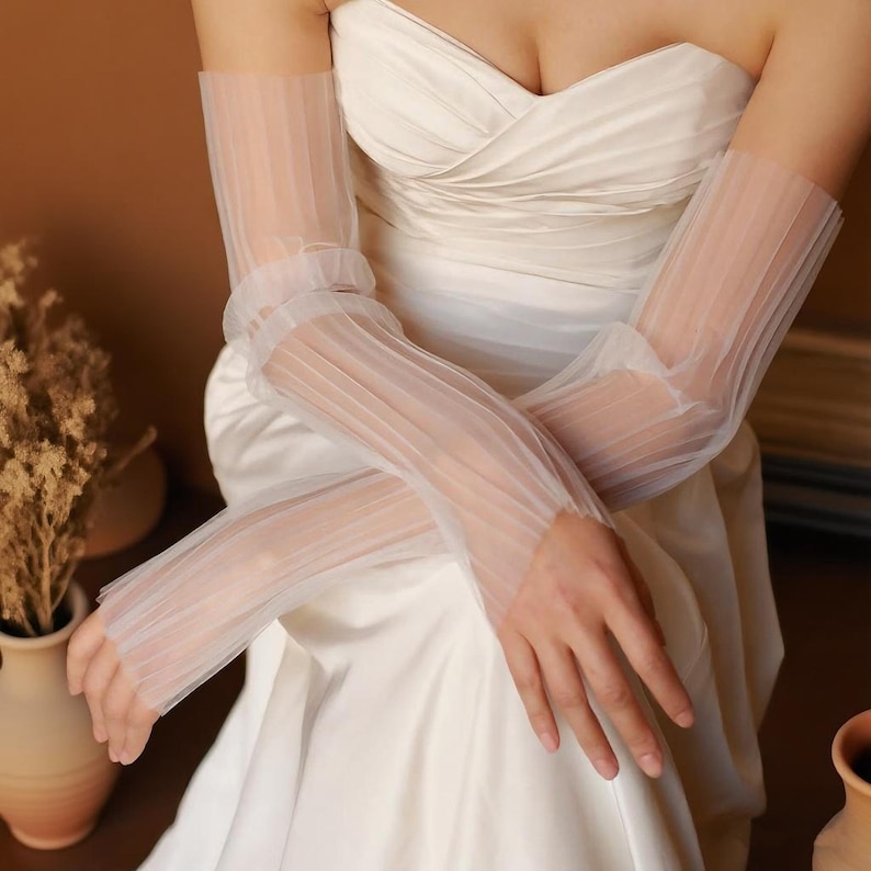 May include: Elegant white sheer gloves with vertical pleats, worn with a white strapless gown. The gloves extend from the fingertips to the upper arm, creating a delicate and sophisticated look. Perfect for a wedding or formal event.