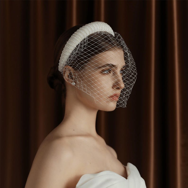 May include: A white bridal headband with a pearl and bead design, paired with a delicate net veil. The headband is thick and padded, and the veil is a fine mesh. The background is a solid brown curtain.