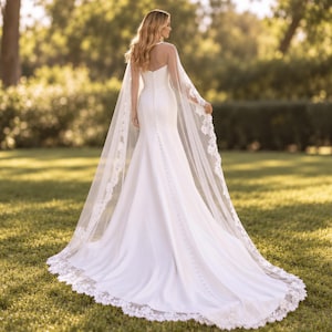 May include: A floor-length white gown with a fitted bodice and a long train. The back features a vertical row of small buttons extending from the neckline to the hem. A sheer, floor-length cape with intricate white lace trim drapes over the shoulders.