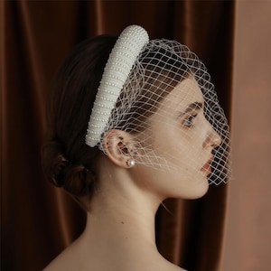 May include: A white bridal headband with pearl embellishments and a delicate net veil. The headband is thick and cylindrical, covered in small, evenly spaced pearls. The veil is a fine, open weave, creating a soft, romantic look.