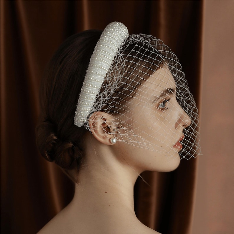 May include: A white bridal headband with pearl embellishments and a delicate net veil. The headband is thick and cylindrical, covered in small, evenly spaced pearls. The veil is a fine, open weave, creating a soft, romantic look.