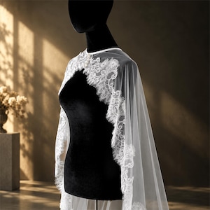 May include: A white bridal capelet featuring intricate floral lace trim along the edges and a sheer tulle fabric. The garment is draped over a black mannequin, showcasing a scalloped lace border and a simple round neckline with a delicate pearl accent.