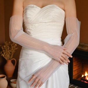 May include: Sheer, white, pleated bridal gloves. The gloves are long, extending from the fingertips to the upper arm. The fabric is delicate and translucent, with a subtle texture. The gloves are worn with a white strapless dress.
