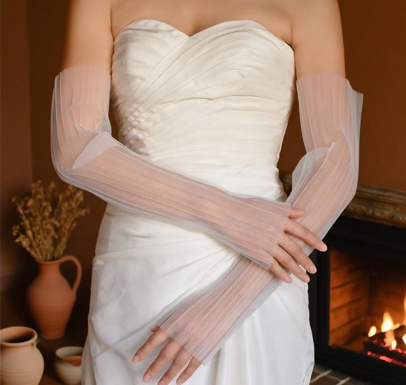 May include: Sheer, white, pleated bridal gloves. The gloves are long, extending from the fingertips to the upper arm. The fabric is delicate and translucent, with a subtle texture. The gloves are worn with a white strapless dress.