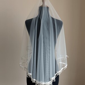 Op de afbeelding: Een witte bruidssluier van een delicate, transparante stof. De sluier is versierd met een wit satijnen lint en kleine, bungelende parelachtige kralen langs de rand. Het wordt getoond op een zwarte mannequin.