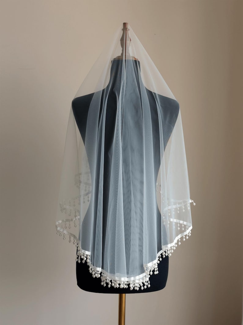 Op de afbeelding: Een witte bruidssluier van een delicate, transparante stof. De sluier is versierd met een wit satijnen lint en kleine, bungelende parelachtige kralen langs de rand. Het wordt getoond op een zwarte mannequin.