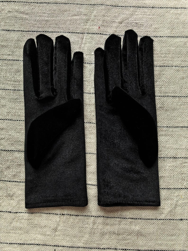 May include: A pair of black velvet gloves featuring a smooth, soft-touch fabric finish. The gloves have a classic five-finger design with a solid black color throughout, showcasing a consistent texture and a clean, straight-cut wrist opening.