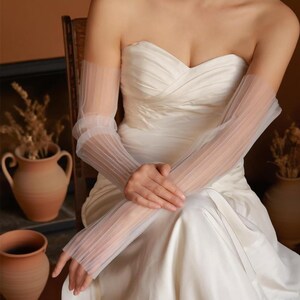 May include: Elegant, sheer white bridal gloves with a pleated design. The gloves are long, extending from the upper arm to the fingers. The gloves are worn with a strapless white wedding dress. The overall aesthetic is delicate and romantic.