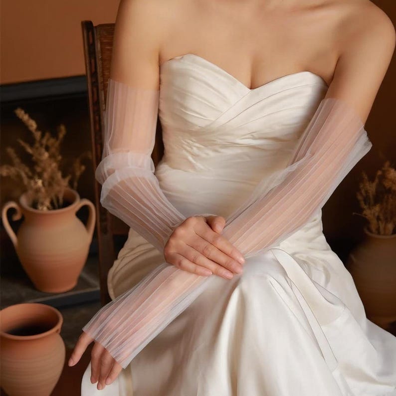 May include: Elegant, sheer white bridal gloves with a pleated design. The gloves are long, extending from the upper arm to the fingers. The gloves are worn with a strapless white wedding dress. The overall aesthetic is delicate and romantic.