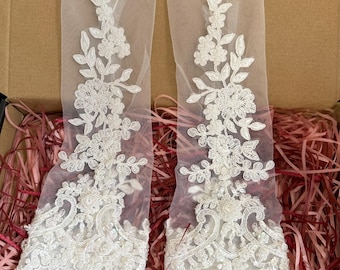 Könnte beinhalten: Ein Paar elfenbeinfarbene, fingerlose Brauthandschuhe mit aufwendigen floralen Spitzendetails. Die Handschuhe sind aus transparentem Stoff gefertigt und werden in einer Pappschachtel mit roter Papierfüllung präsentiert. Der Text "Vow Tulle" ist sichtbar.
