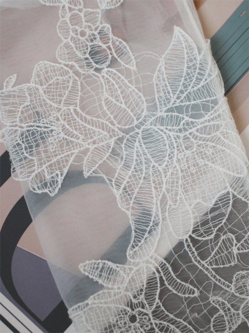 May include: Close-up of white floral lace fabric. The lace features intricate, detailed designs of flowers and leaves. The fabric is semi-sheer, with a delicate, airy appearance. The background is a mix of soft colors.