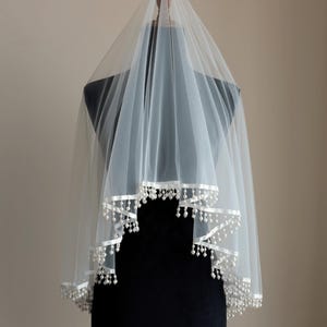 Op de afbeelding: Een witte bruidssluier van transparante stof. De rand is afgewerkt met een wit lint en kleine, parelachtige kralen. De sluier wordt gepresenteerd op een zwarte mannequin. Het ontwerp is elegant en eenvoudig.