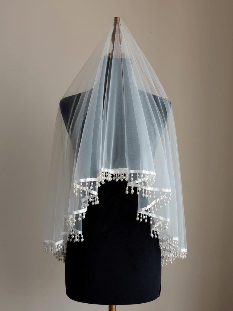 Op de afbeelding: Een witte bruidssluier van transparante stof. De rand is afgewerkt met een wit lint en kleine, parelachtige kralen. De sluier wordt gepresenteerd op een zwarte mannequin. Het ontwerp is elegant en eenvoudig.