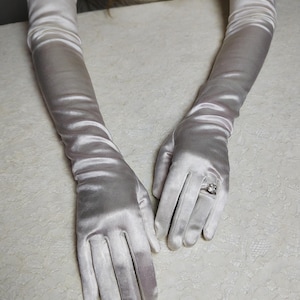 Könnte beinhalten: Ein Paar lange Satinhandschuhe in einem weichen, silbrig-weißen Farbton. Die Handschuhe reichen über die Ellbogen hinaus und sind am Oberarm gerafft. Ein Ring mit einem quadratischen Stein ist an einer Hand zu sehen. Die Handschuhe liegen auf einer weißen, strukturierten Oberfläche.