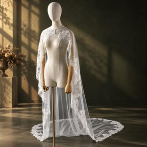 May include: A white bridal cape made of sheer tulle fabric featuring intricate floral lace embroidery along the edges and neckline. The cape drapes over the shoulders and extends to the floor, creating a long train with a scalloped lace border.