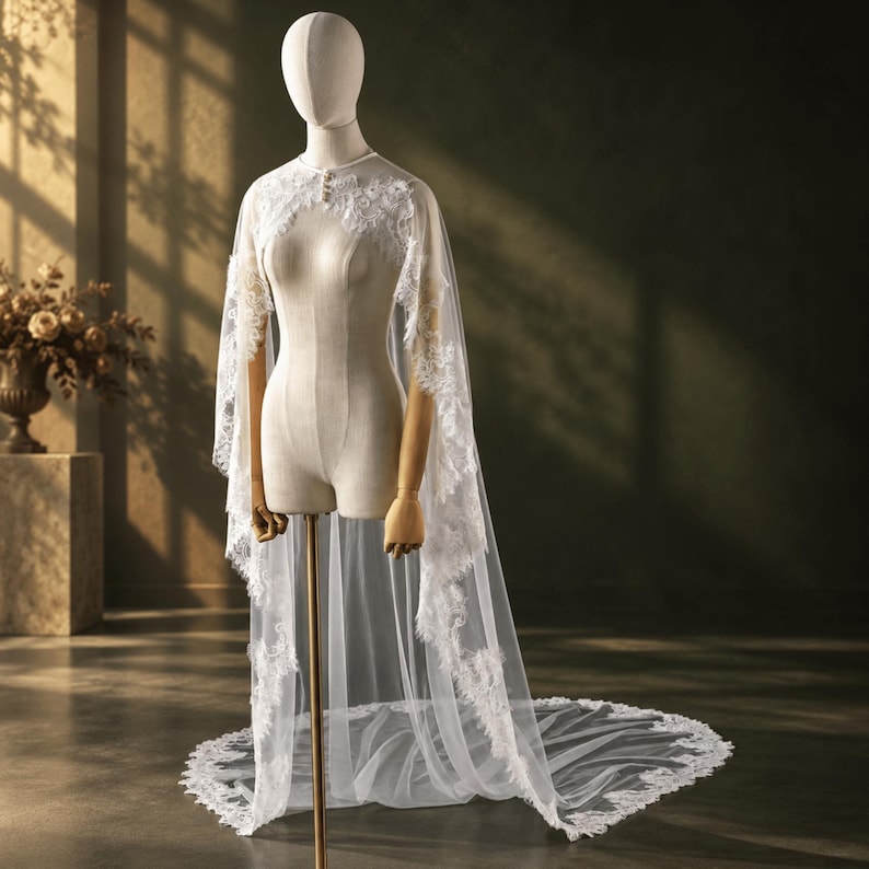 May include: A white bridal cape made of sheer tulle fabric featuring intricate floral lace embroidery along the edges and neckline. The cape drapes over the shoulders and extends to the floor, creating a long train with a scalloped lace border.